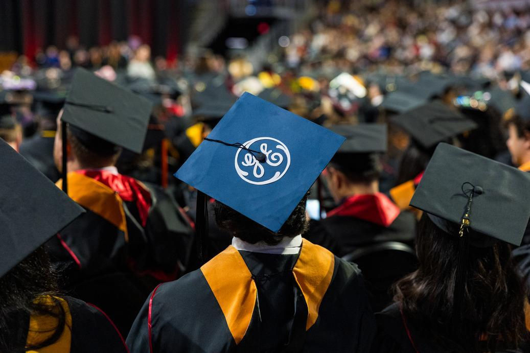 the back of many graduating students in cap and gown with one student's cap emblazoned with GE on the cap