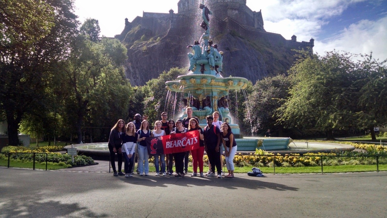Students posing with a UC flag in front of a fountain in Scotland.