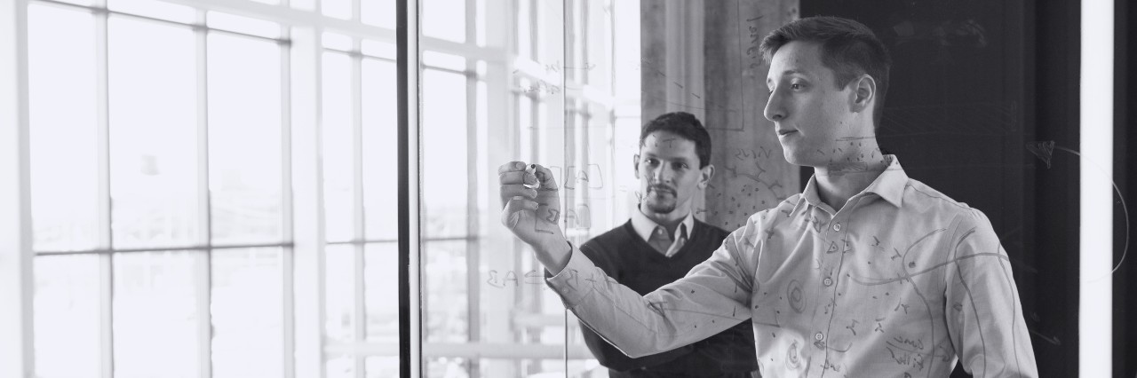 Two students writing on a glass whiteboard 